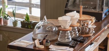 Pour-over drippers and a glass carafe on the shop's wooden bar, with plants along the window behind