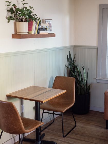 A corner table with two leather chairs, a wall shelf of books, and a plant, under string lights
